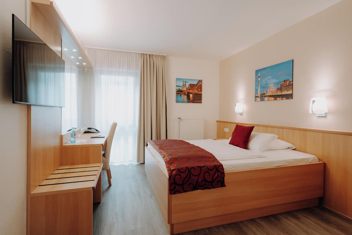 Quarto de hotel com cama, escrivaninha, cadeira, televisão e duas luminárias de parede.