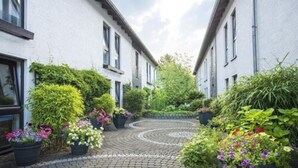 A narrow courtyard with a cobblestone path, surrounded by white buildings with multiple windows and lush greenery including potted plants and trees. - Hotel Am Wald (Monheim)