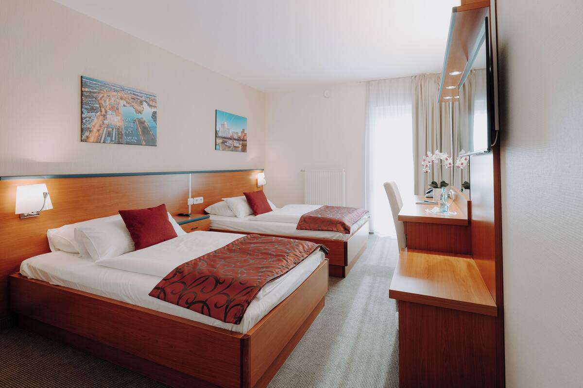 Quarto de hotel com duas camas, uma escrivaninha, uma cadeira, televisão e obras de arte nas paredes.