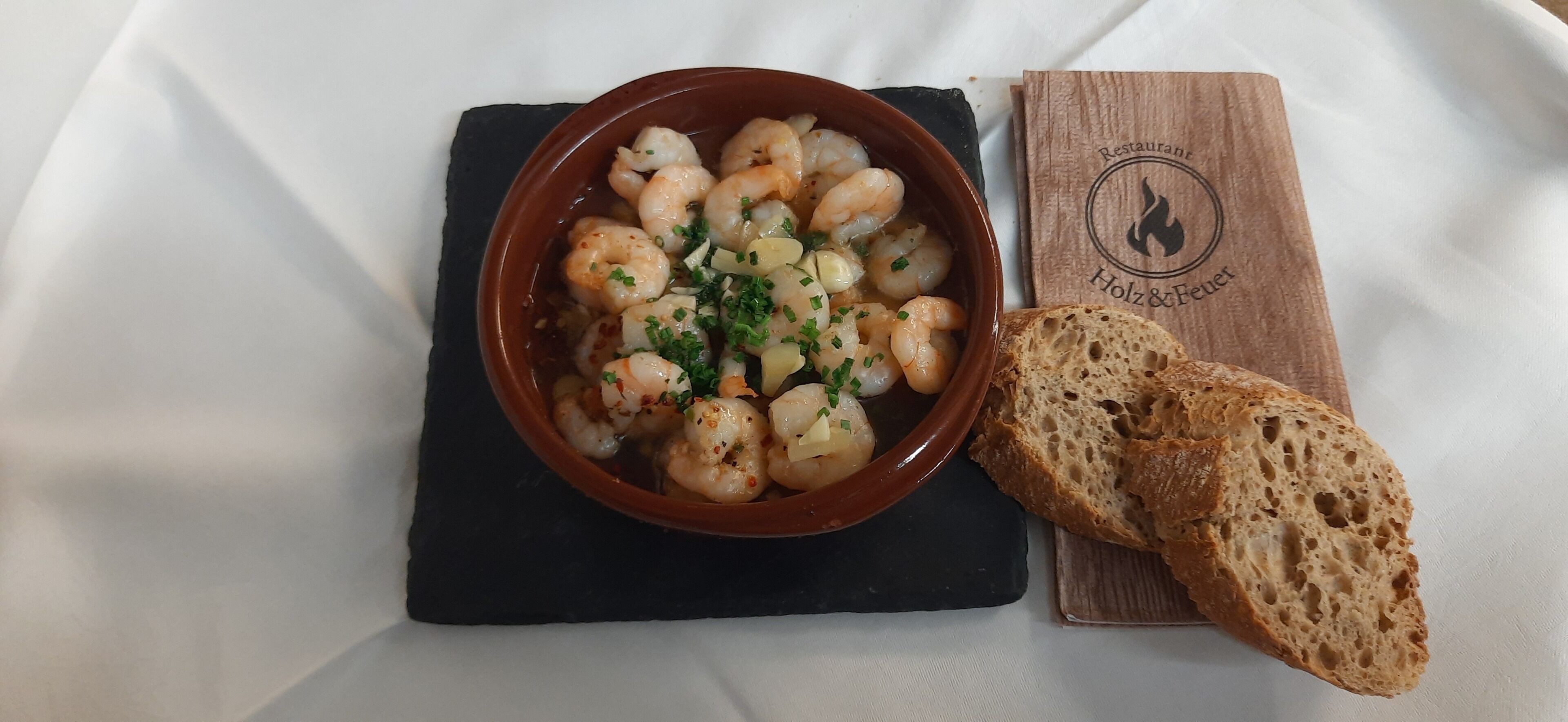 A bowl of shrimp dish with bread on a black slate, placed on a white tablecloth.