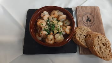A bowl of shrimp dish with bread on a black slate, placed on a white tablecloth.