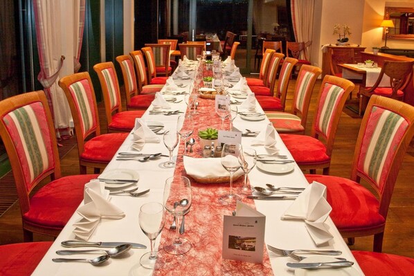 Una larga mesa de comedor dispuesta para una comida, con sillas tapizadas en rojo, un mantel blanco y un camino de mesa rojo estampado.