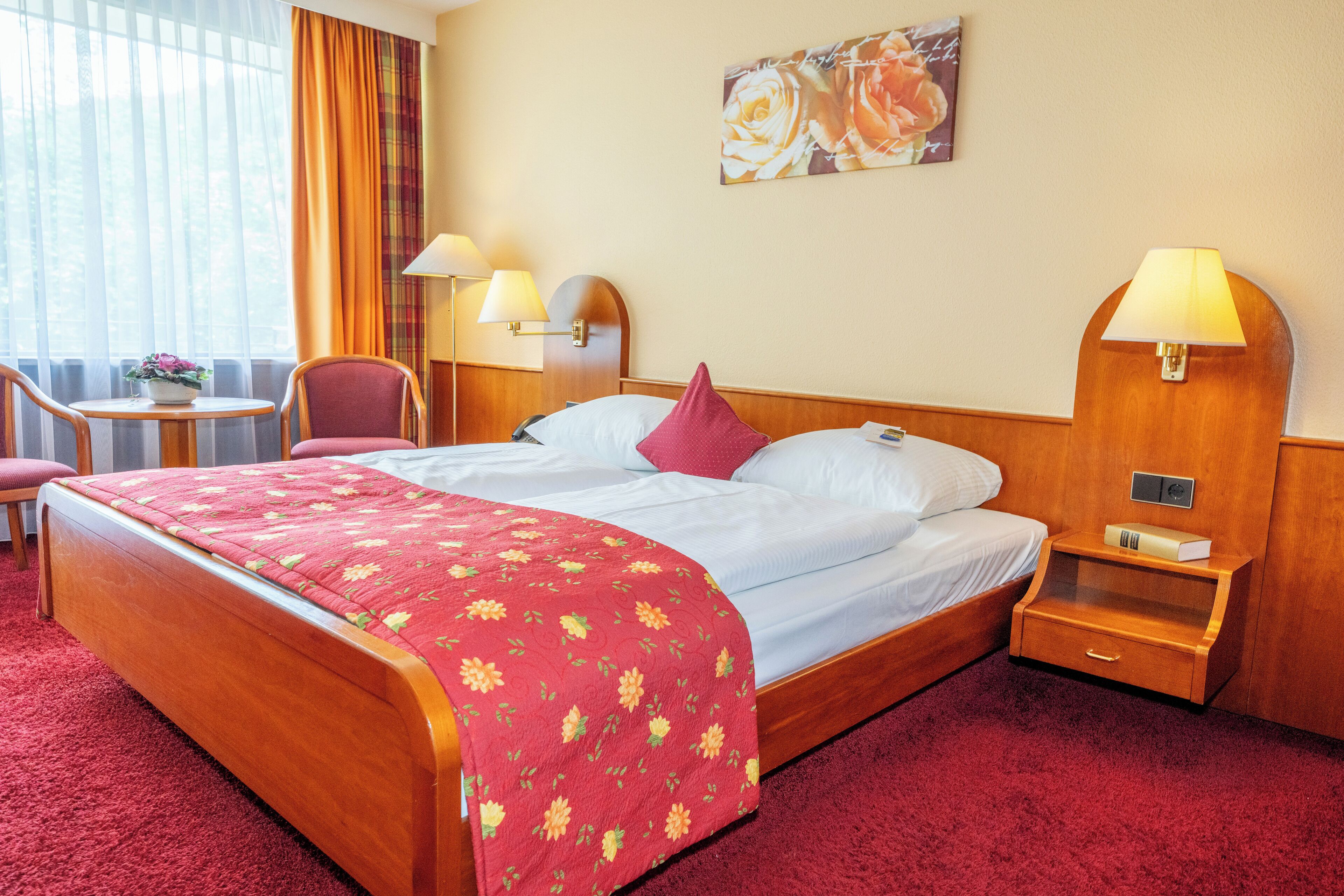 A hotel room with a large bed, a bedside table with a lamp, a small table with a chair, and a painting on the wall.