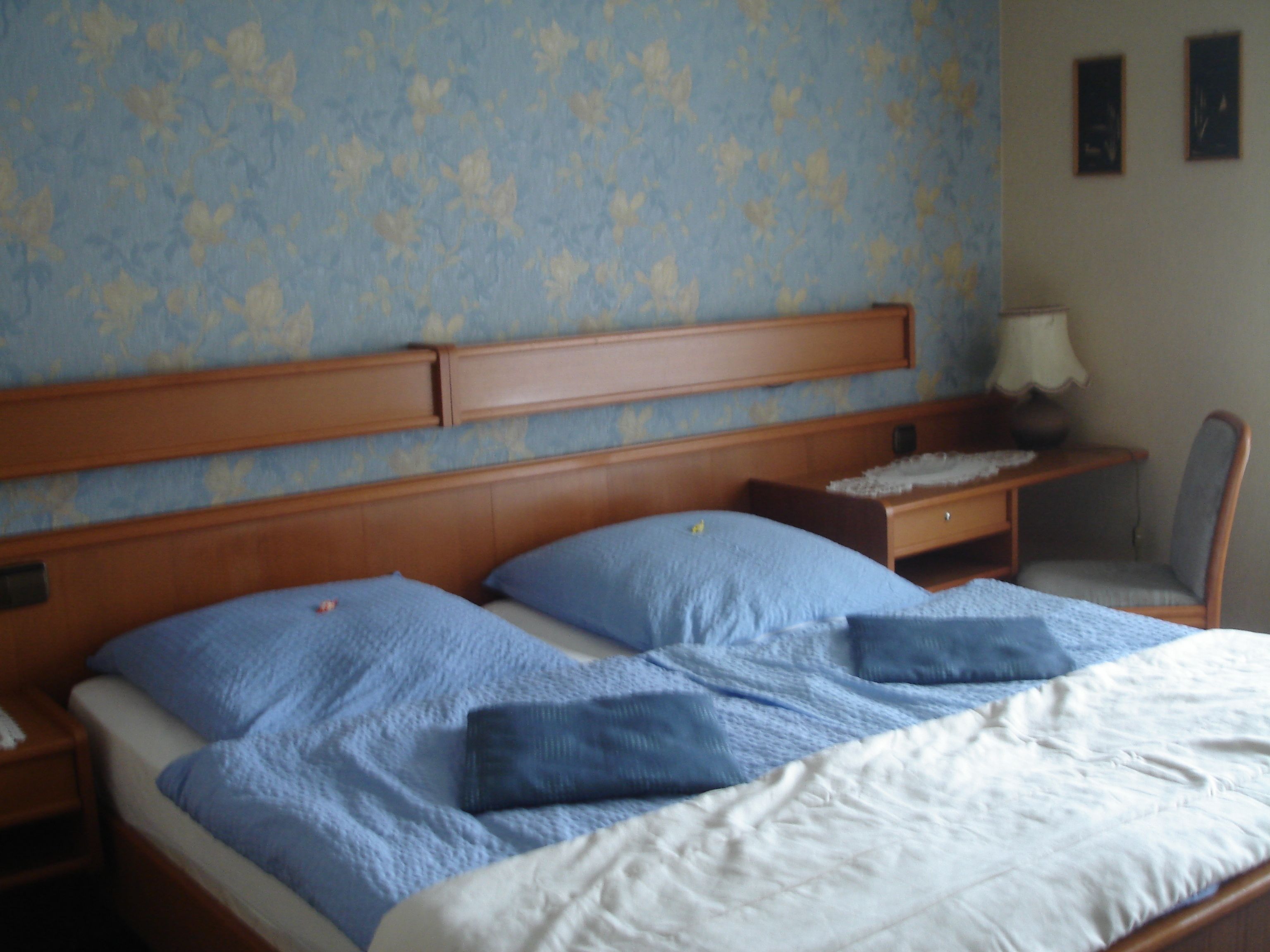 A double bed with blue and white bedding, a wooden headboard, a nightstand with a lamp, and a chair.