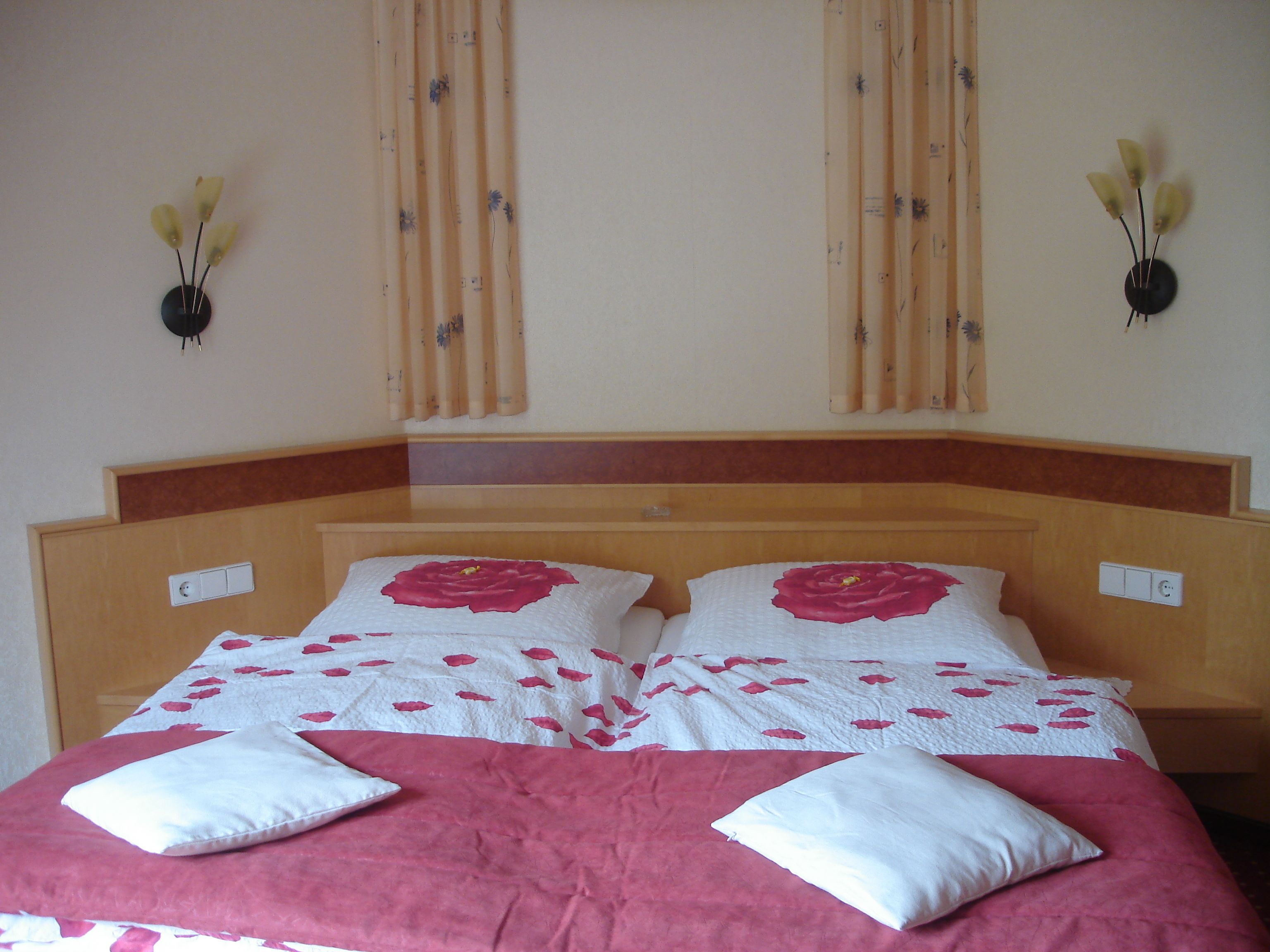 Un lit recouvert d’une couvre-lit à motifs floraux rouges et blancs, avec deux oreillers blancs et une tête de lit en bois.