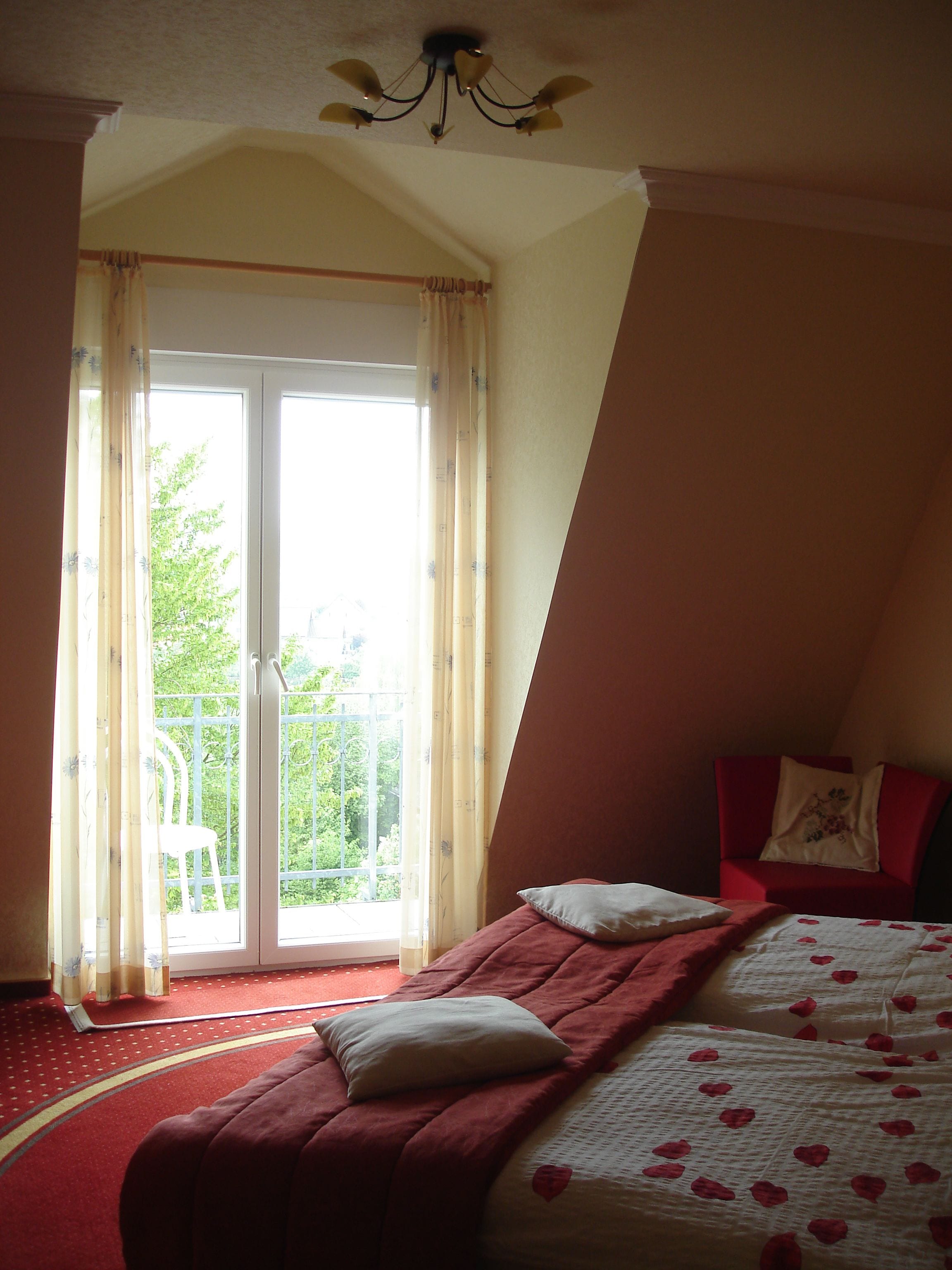 A bedroom with a bed, a red chair, a balcony with a white chair, and a view of greenery outside.