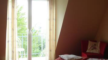 A bedroom with a bed, a red chair, a balcony with a white chair, and a view of greenery outside.