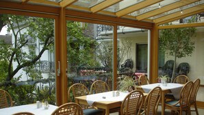 A restaurant with wooden tables and chairs, large windows, and a view of trees and buildings. - Hotel Zum Rehberg (Kastellaun)