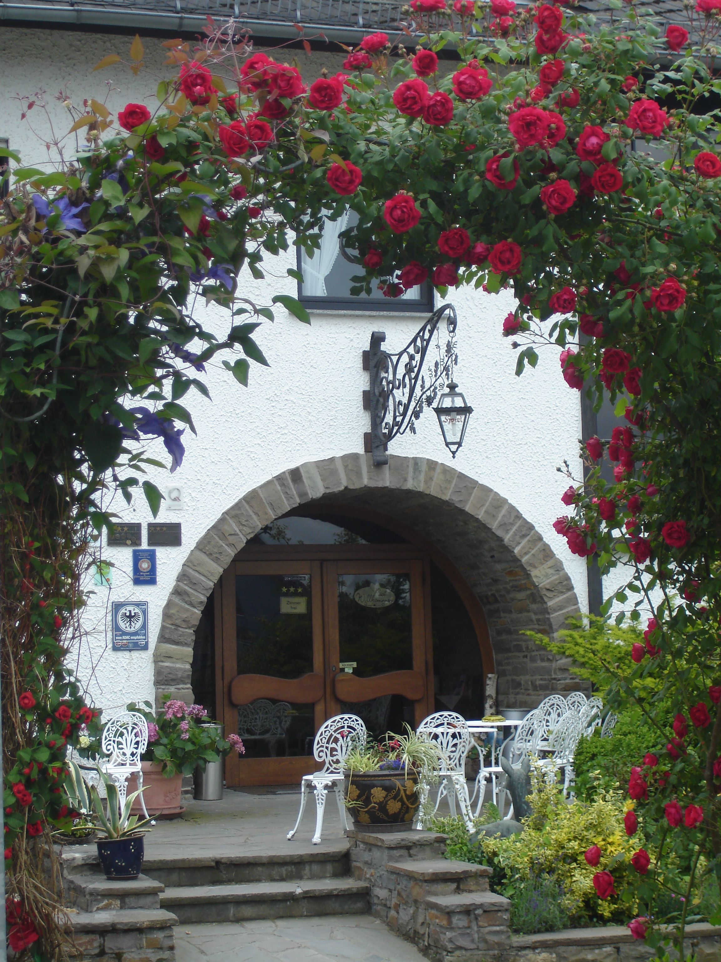 L’entrée d’un bâtiment est ornée d’une arche en pierre, d’une porte en bois et d’une enseigne placée au-dessus de la porte. Des plantes en pot et des chaises blanches se trouvent à l’extérieur.