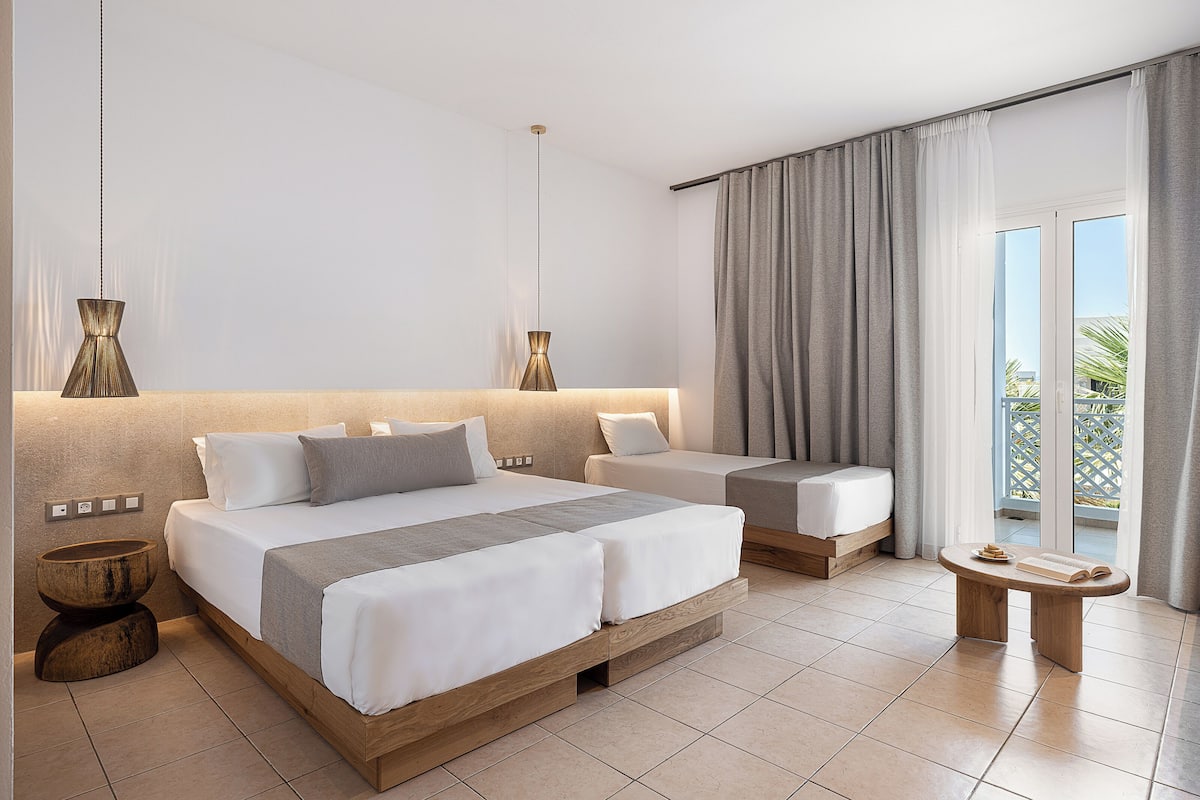 Una habitación de hotel moderna con una cama grande, una cama más pequeña, un banco de madera y vistas al exterior.