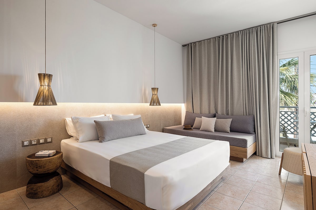 Una habitación de hotel moderna con una cama grande, un sofá más pequeño, una mesita de madera y vistas al exterior a través de un ventanal corredero.