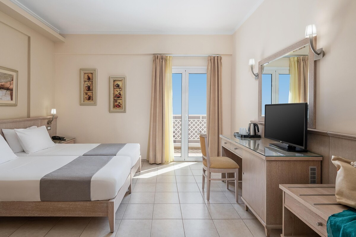 A hotel room with two beds, a desk with a television, a chair, and a balcony with curtains.