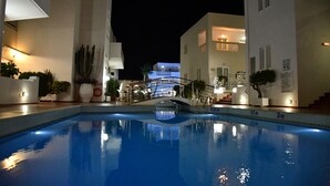 Zona de piscina nocturna con vista despejada de la piscina, un puente y edificios circundantes.
