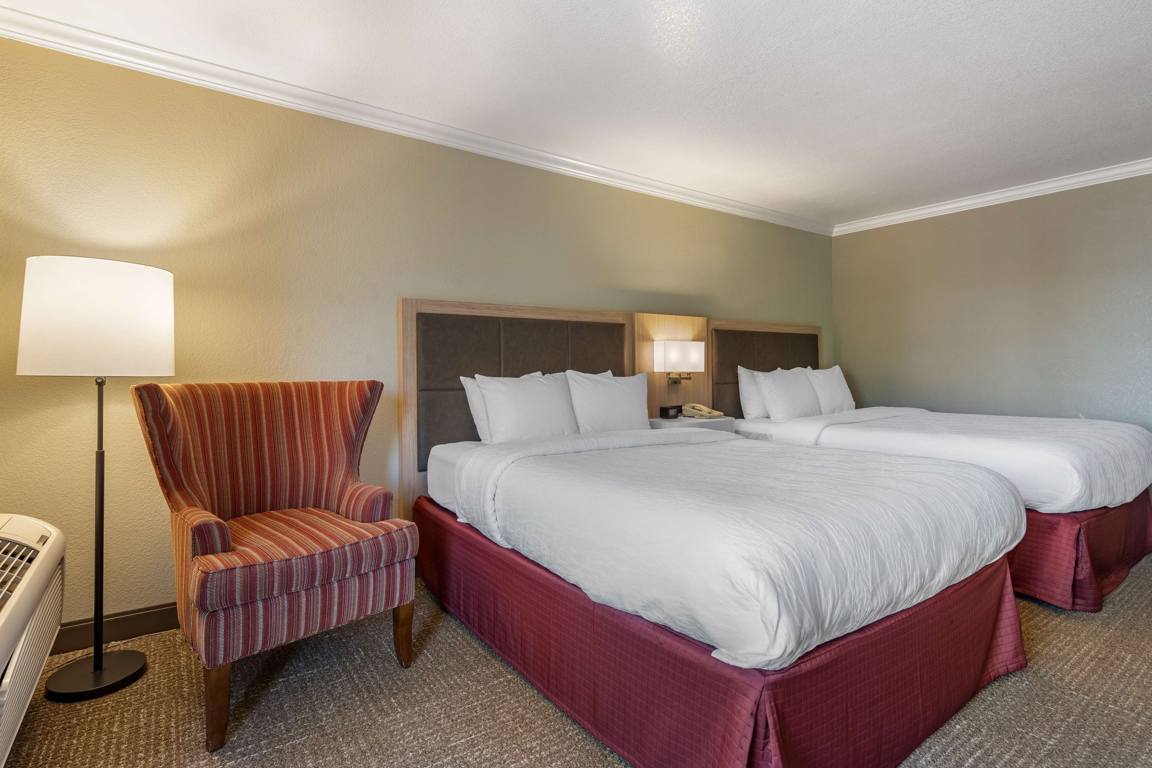 Une chambre d’hôtel avec deux lits, un fauteuil à rayures, une lampe sur pied et un radiateur.
