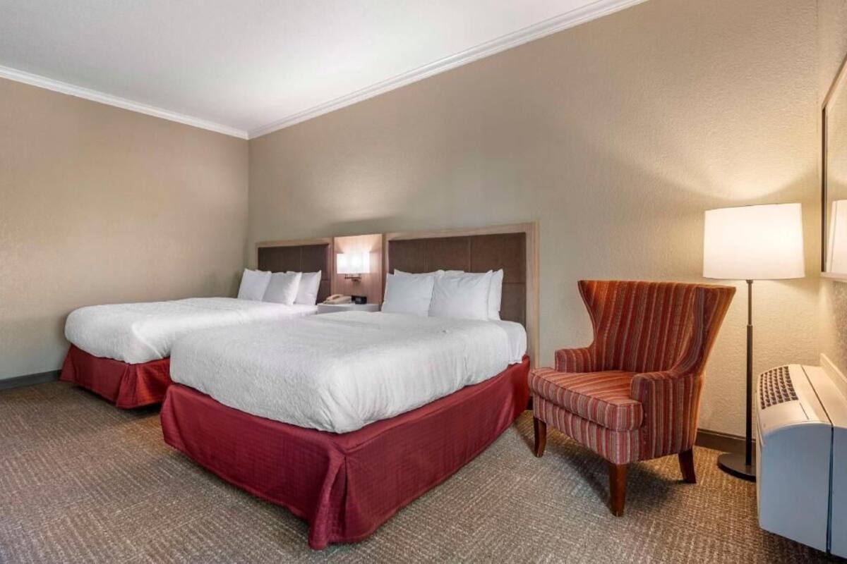 A hotel room with two beds, a chair, a lamp, and a desk with a computer.