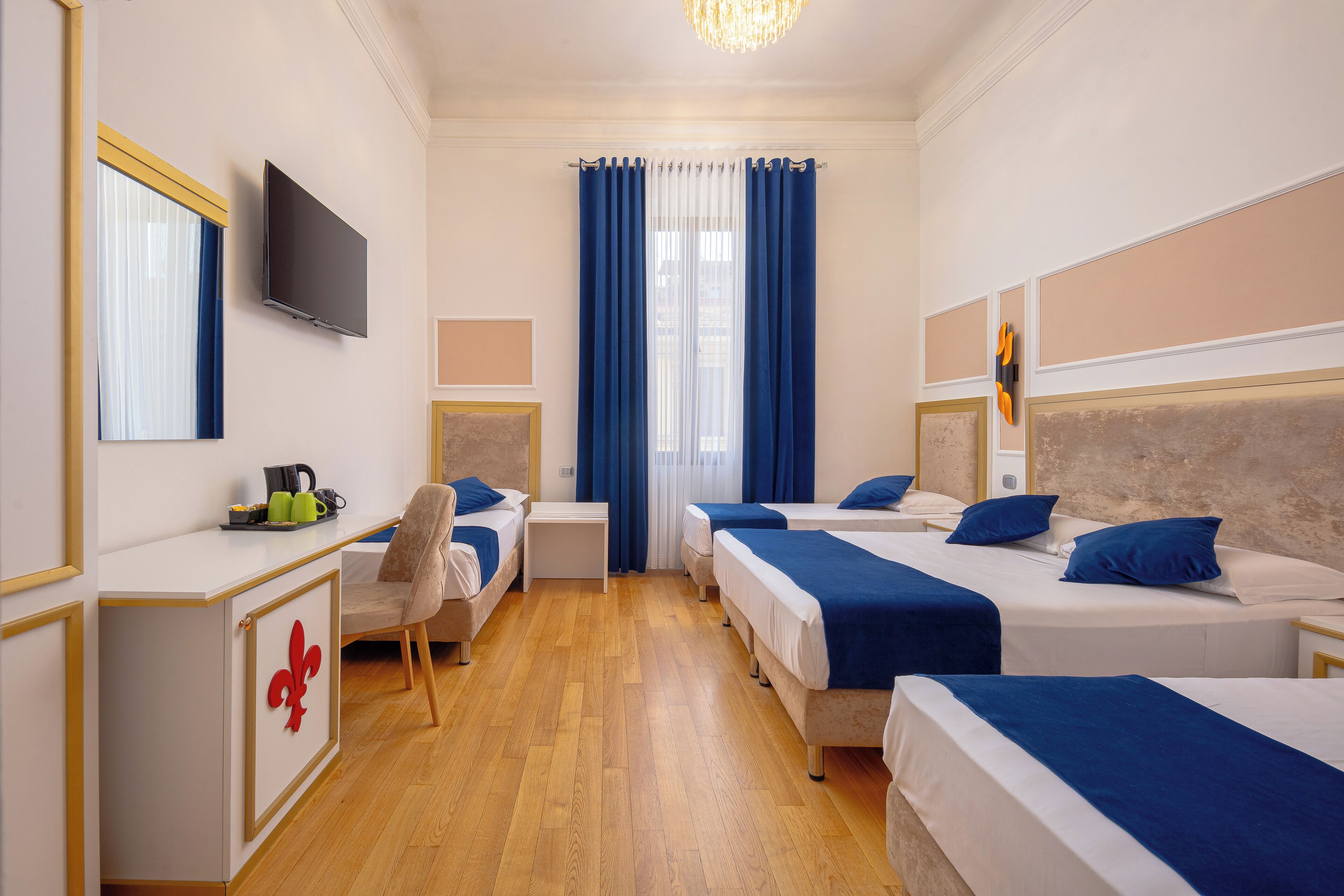 A hotel room with four beds, a desk with a chair, a television, and a large window with blue curtains.