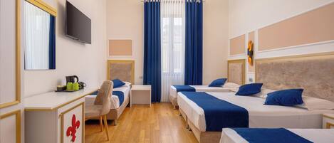 A hotel room with four beds, a desk with a chair, a television, and a large window with blue curtains.
