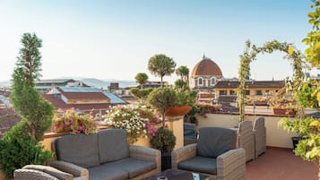 Terraza en la azotea con mobiliario de mimbre, plantas en macetas y vista a un paisaje urbano que incluye un edificio con cúpula.