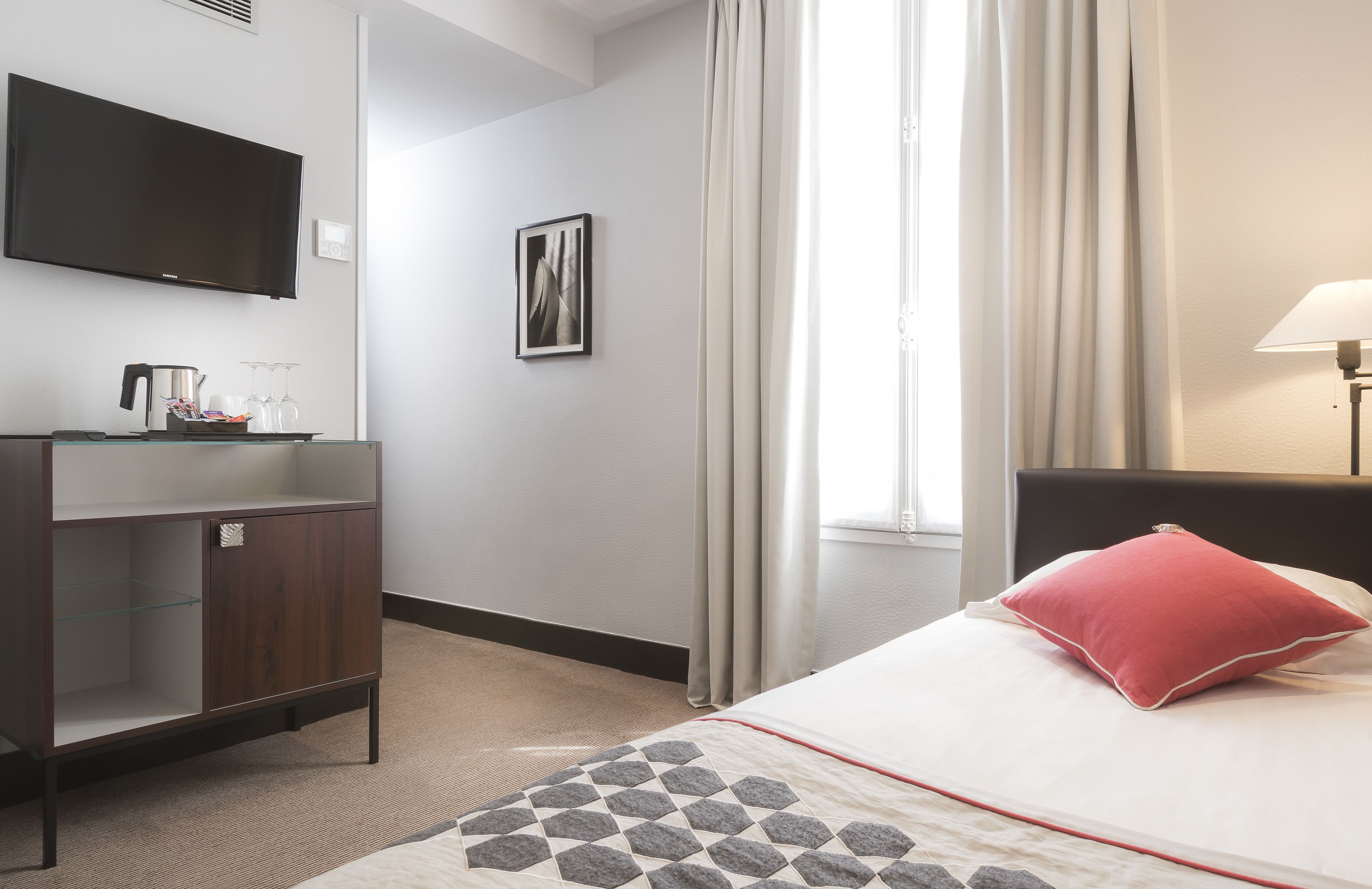 A hotel room with a bed, a TV mounted on the wall, a bedside table with a lamp, a picture on the wall, and a window with curtains.