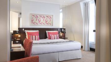 A hotel room with a large bed, two bedside tables with lamps, a red chair, a large window with curtains, and a framed abstract artwork on the wall.