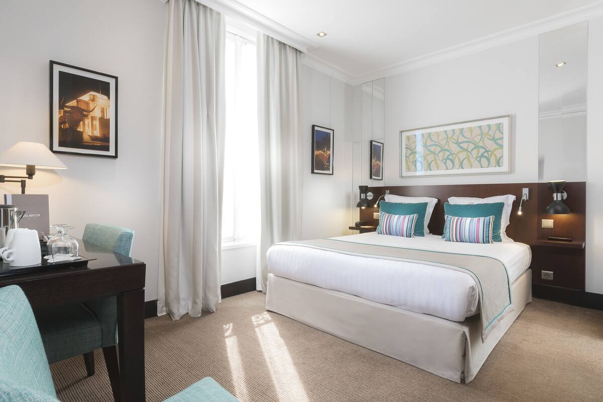 Une chambre d’hôtel avec un grand lit, un bureau avec une lampe et une chaise. Il y a des rideaux à la fenêtre et des œuvres d’art accrochées aux murs.
