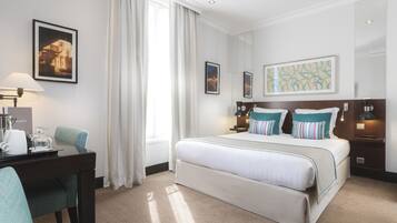 A hotel room with a large bed, a desk with a lamp, and a chair. There are curtains on the window, and artwork on the walls.