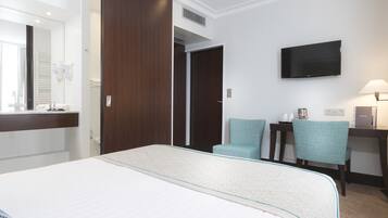 Habitación de hotel con cama, televisor, dos sillas, escritorio con lámpara y espejo.