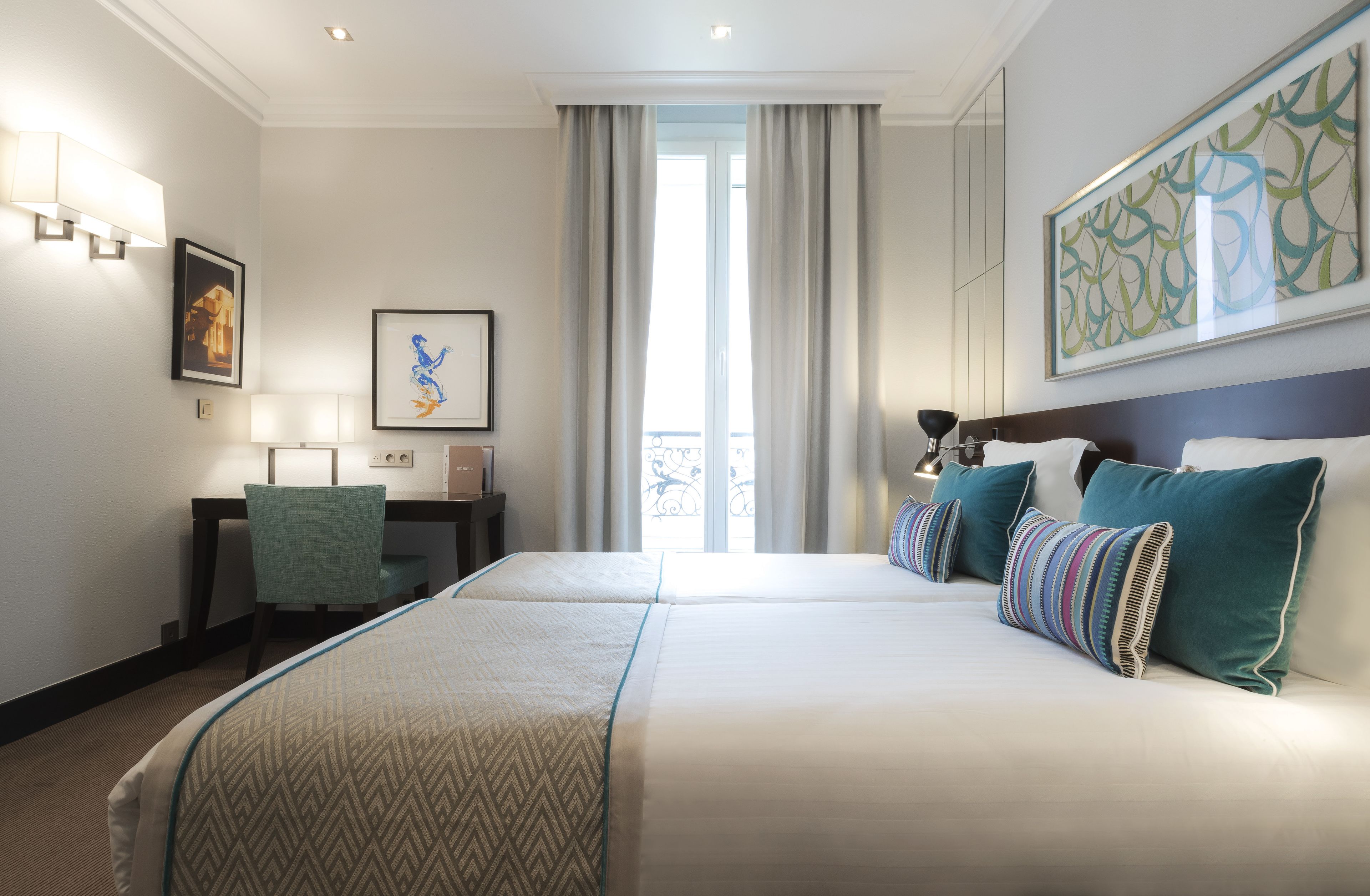 A hotel room with a large bed, a desk with a chair, a window with curtains, and artwork on the walls.