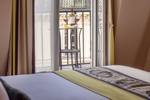 A hotel room with a bed, a window with curtains, a balcony with a table and chairs, and a bottle of wine on the balcony.