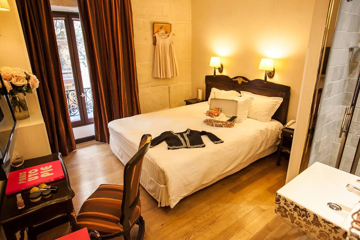 A hotel room with a bed, bedside lamps, a desk with a laptop, a chair, and a bathroom with a sink and mirror.