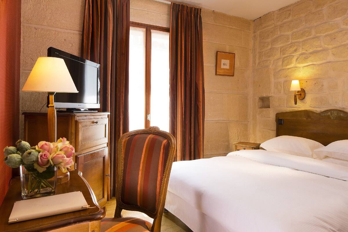 A hotel room with a bed, a television on a wooden cabinet, a chair, a desk with a lamp, and a vase of flowers.