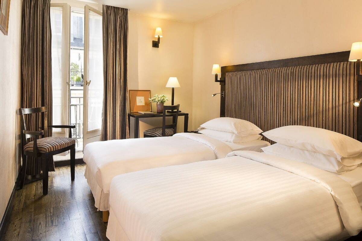 Ein Hotelzimmer mit zwei Betten, einem Sessel, einem kleinen Tisch mit Lampe und Blick ins Freie durch eine Tür.