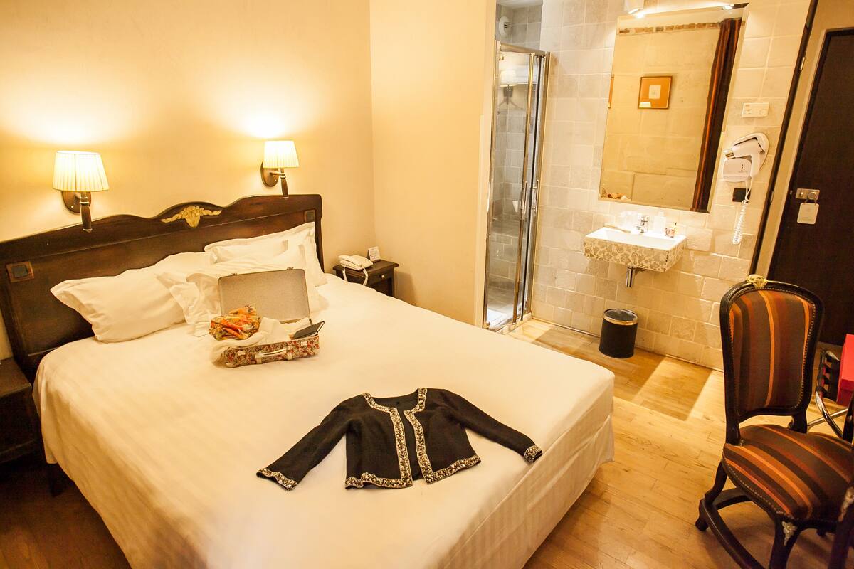 A hotel room with a neatly made bed, a bedside table with a lamp, a shower area, a sink, and a chair.