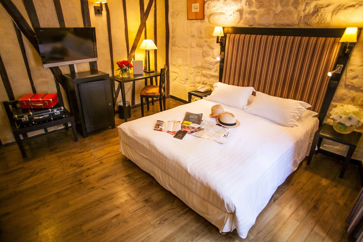 A hotel room with a large bed, a desk with a lamp, a television, and a stone wall.