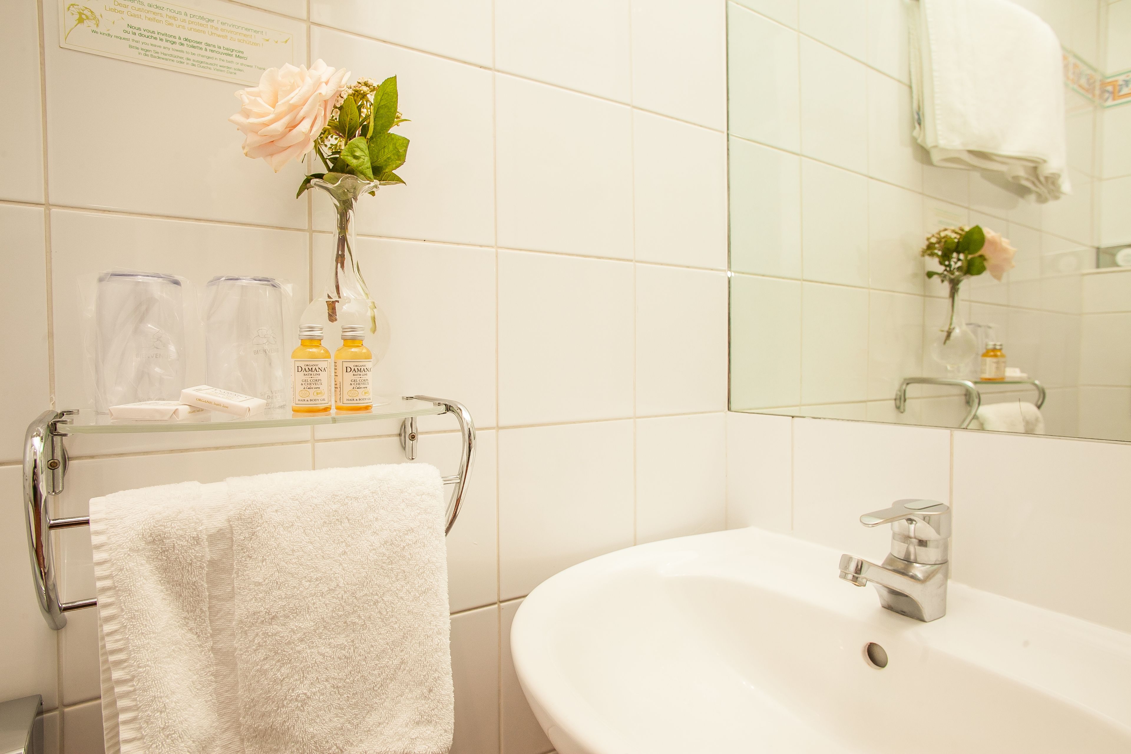 A bathroom with a white sink, a glass shelf with toiletries, a towel rack with a white towel, and a mirror reflecting a rose.
