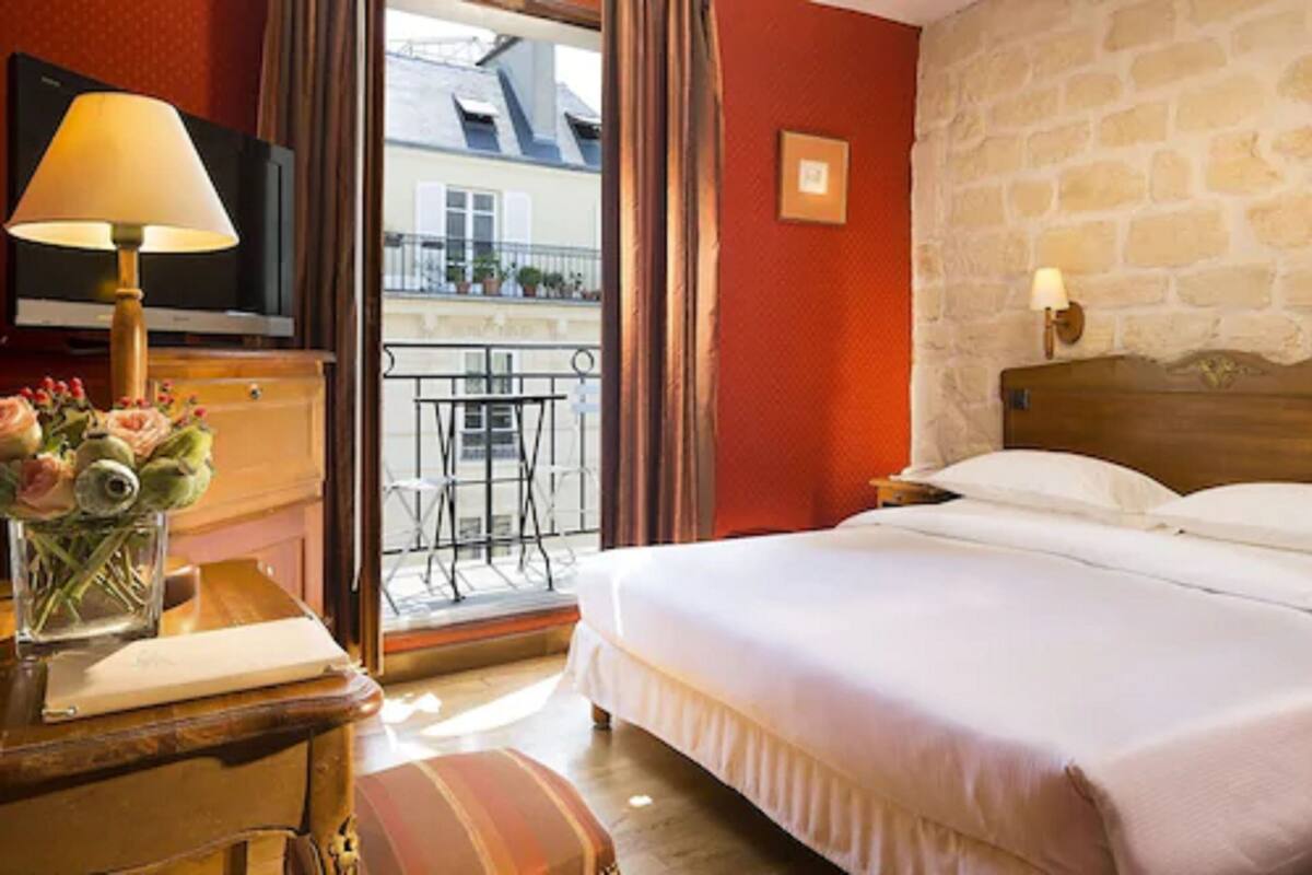 A hotel room with a bed, a desk with a lamp, a TV, and a view of the outside balcony.