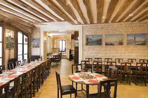 Una sala ristorante con lunghe tavole apparecchiate, travi in legno a soffitto e pareti in pietra con quadri appesi.