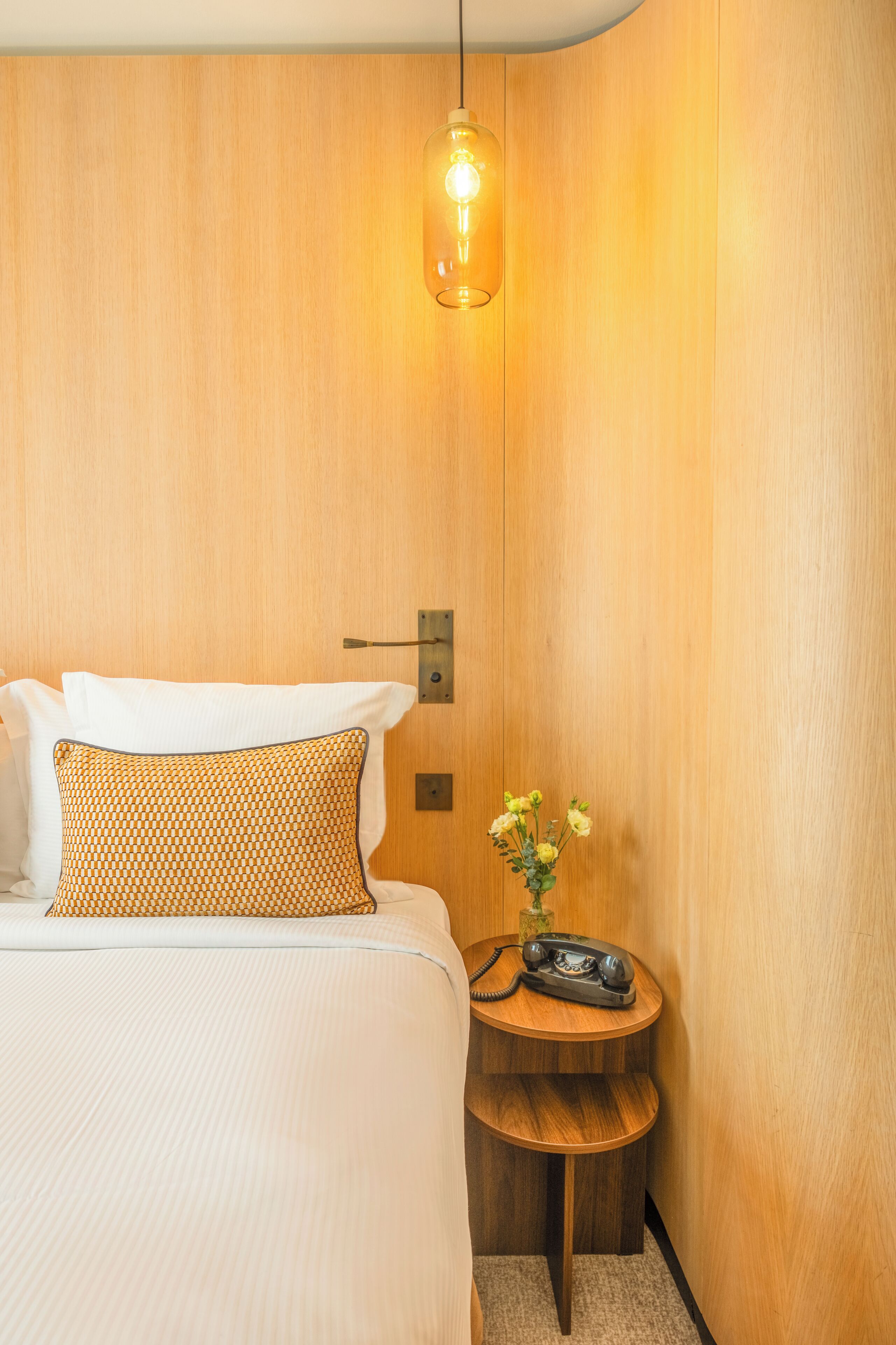 Ein Hotelzimmer mit einer Holzwand, einem Bett mit weißer Bettwäsche, einem gemusterten Kissen, einem kleinen Nachttisch mit Blumen in einer Vase und einer hängenden Lampenleuchte.