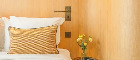 A hotel room with a wooden wall, a bed with white bedding, a patterned pillow, a small wooden side table with a vase of flowers, and a hanging light fixture.