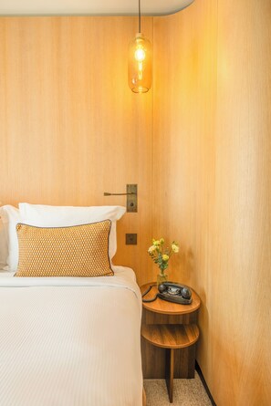 Una camera d'albergo con una parete in legno, un letto con biancheria bianca, un cuscino decorato, un piccolo comodino in legno con un vaso di fiori e un lampadario.