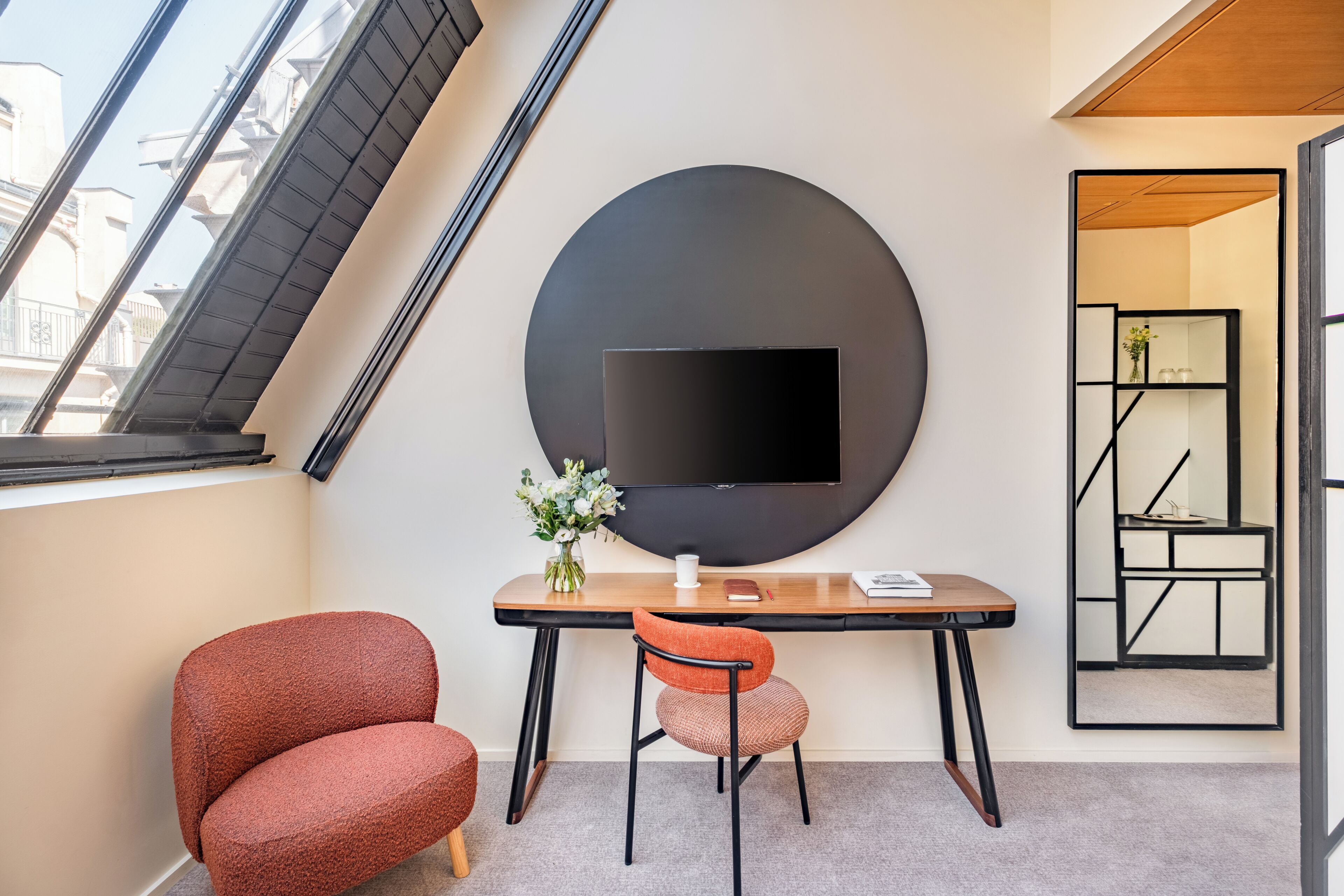 A modern room with a large round wall-mounted TV, a wooden desk, a red chair, and a small table with a vase of flowers.