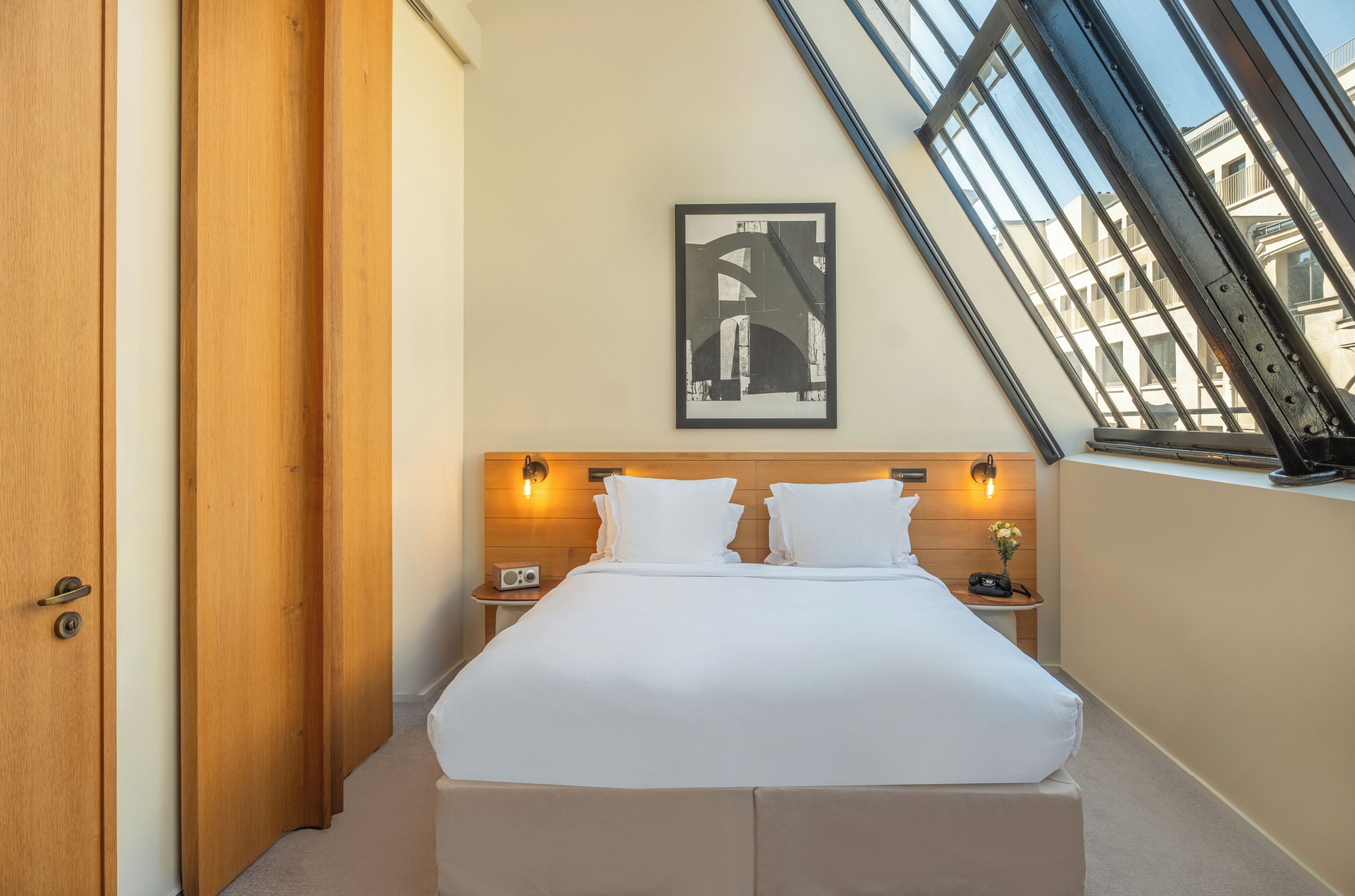 A hotel room with a bed, two bedside tables, a framed picture on the wall, and a window with a view of buildings.