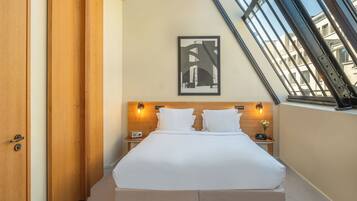 Ein Hotelzimmer mit einem Bett, zwei Nachttischen, einem gerahmten Bild an der Wand und einem Fenster mit Blick auf Gebäude.
