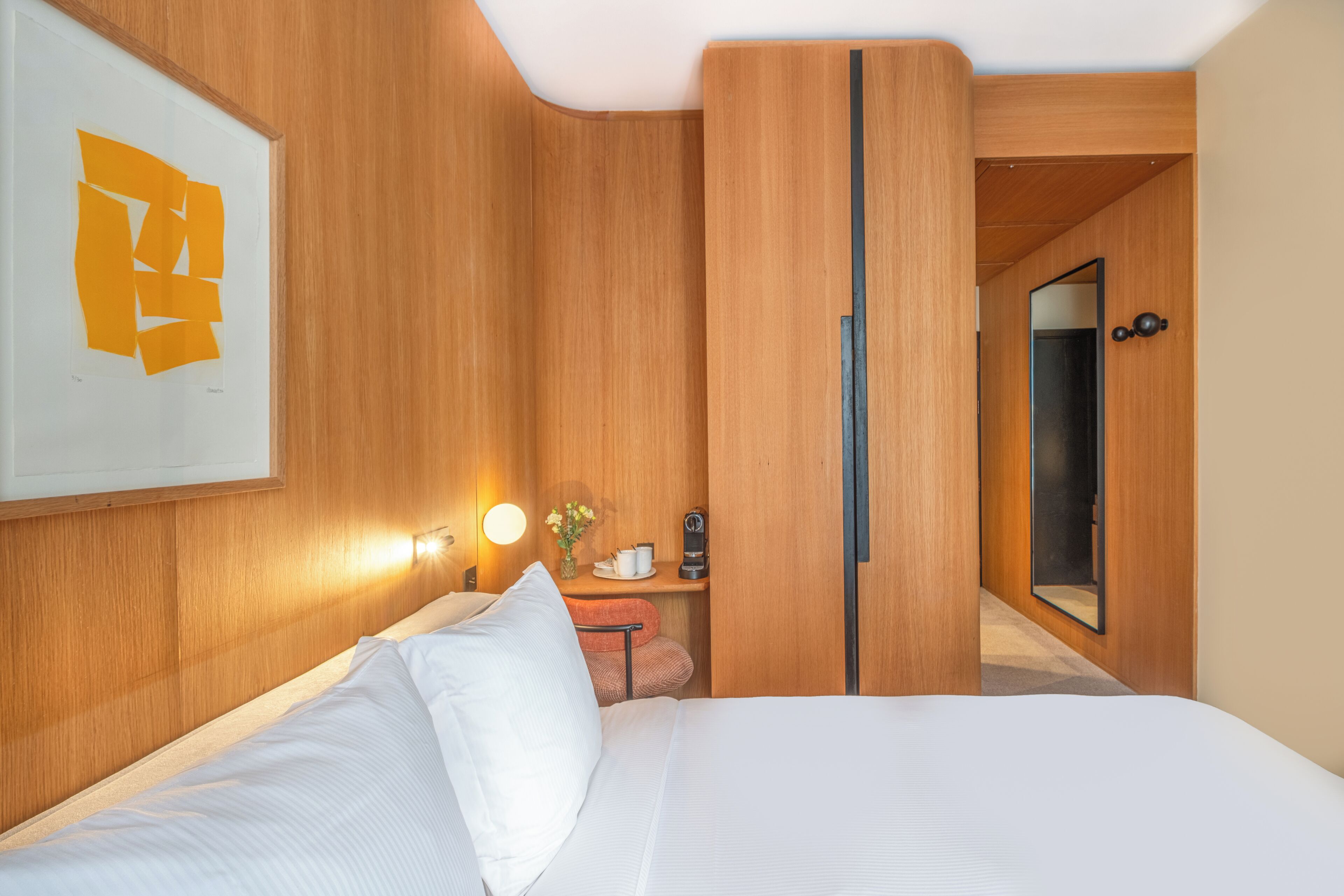 A hotel room with a bed, a nightstand, a mirror, and a wall-mounted light fixture.