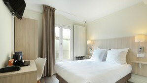 A hotel room with a large bed, a desk with a telephone and a coffee maker, a chair, and a window with curtains.