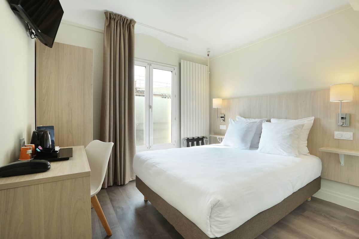 A hotel room with a large bed, a desk with a telephone and a coffee maker, a chair, and a window with curtains.