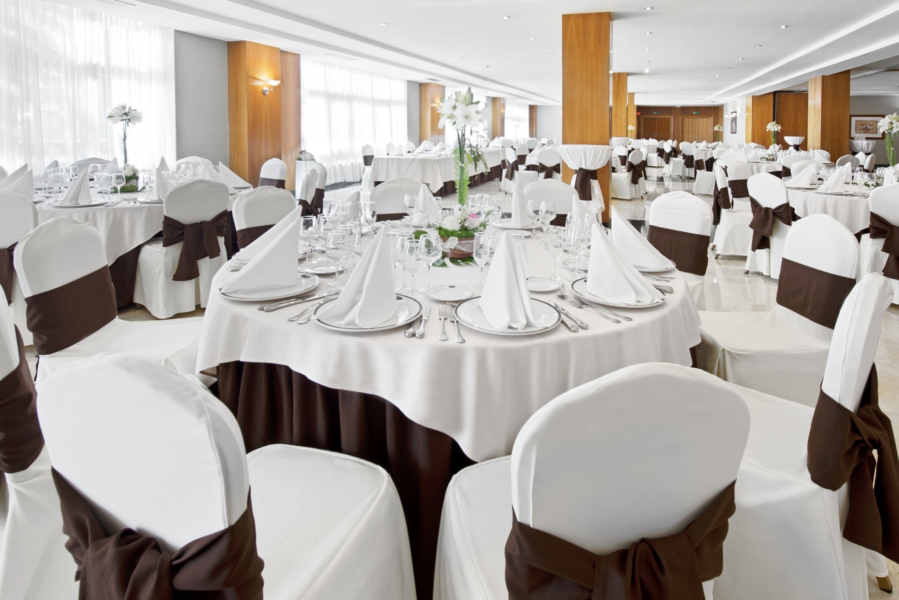 Un salón de banquetes con mesas redondas dispuestas para comer, sillas blancas con cintas marrones y centros de mesa florales.