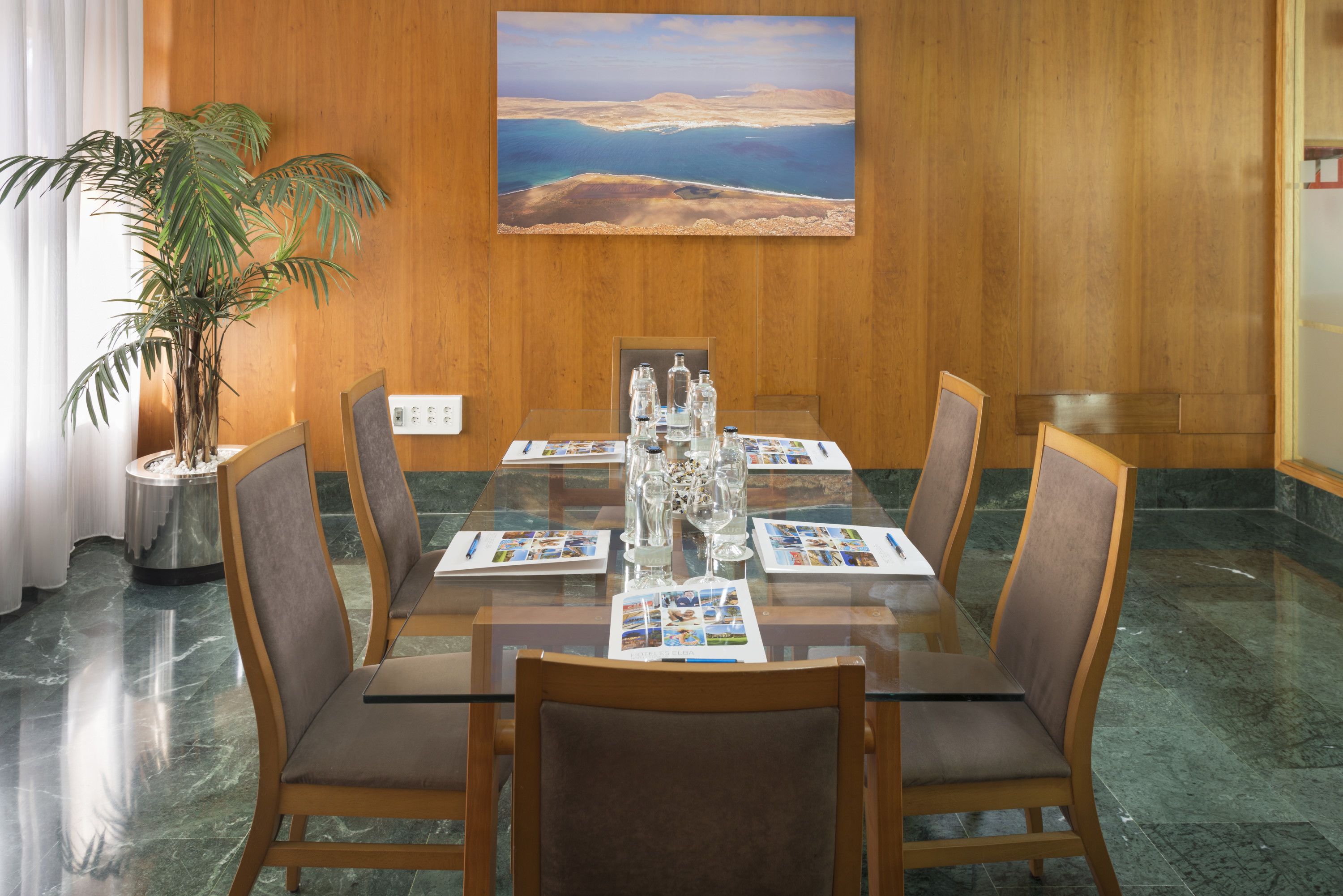 A conference room with a glass table, six chairs, a potted plant, and a painting on the wall.