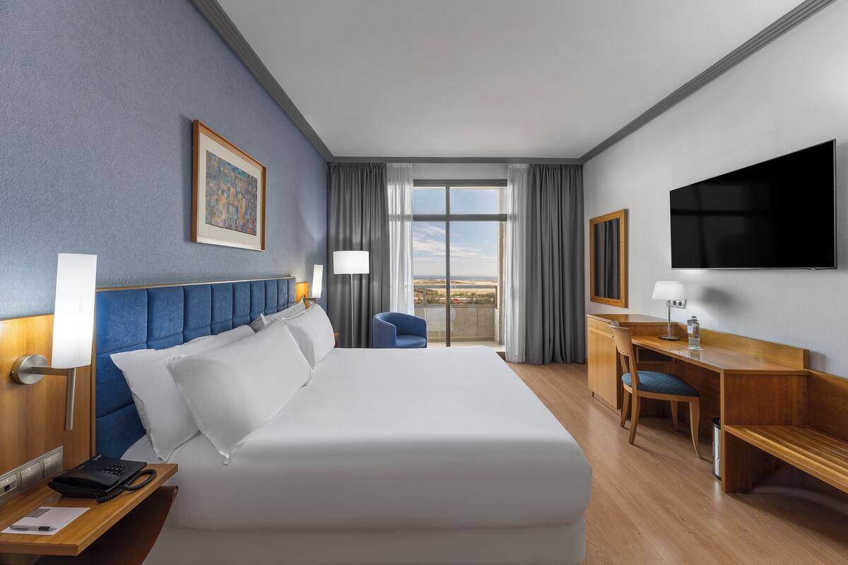 Une chambre d’hôtel avec un grand lit, un bureau, une chaise, une télévision et une fenêtre avec des rideaux.