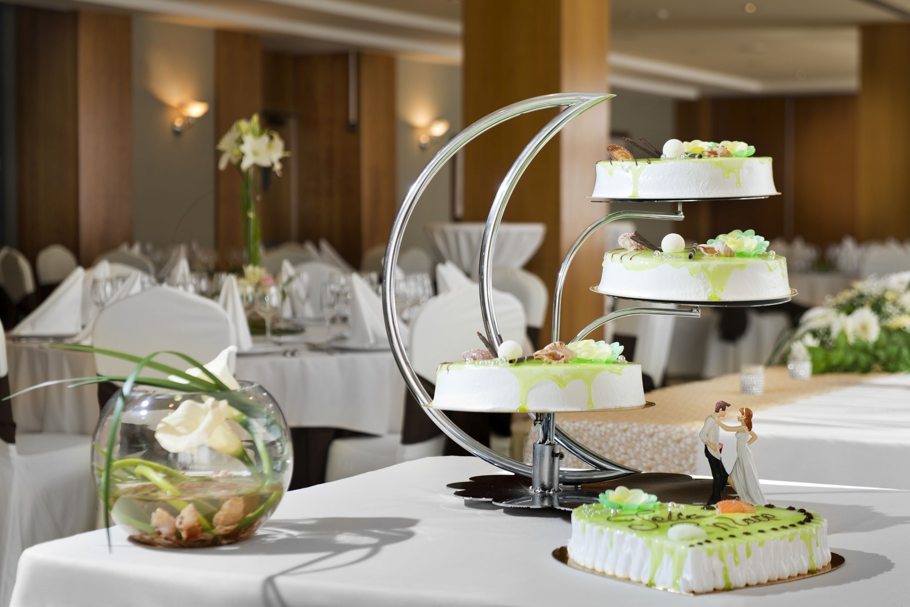 A three-tiered wedding cake with green icing and white decorations on a table in a banquet hall.